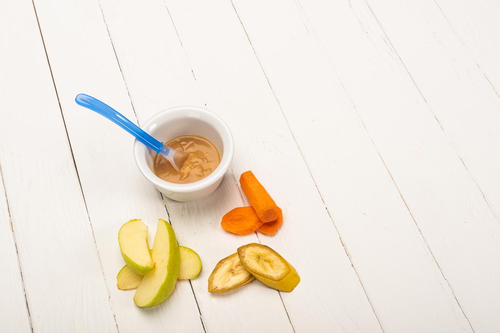 High angle view of baby food in bowl with spoon and sliced fruits and carrot on white wooden surface - Photo, Image