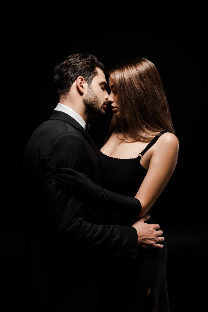 πλαϊνή άποψη του όμορφος γενειοφόρος άνδρας και η γυναίκα φιλιά απομονώνονται σε μαύρο  - Φωτογραφία, εικόνα