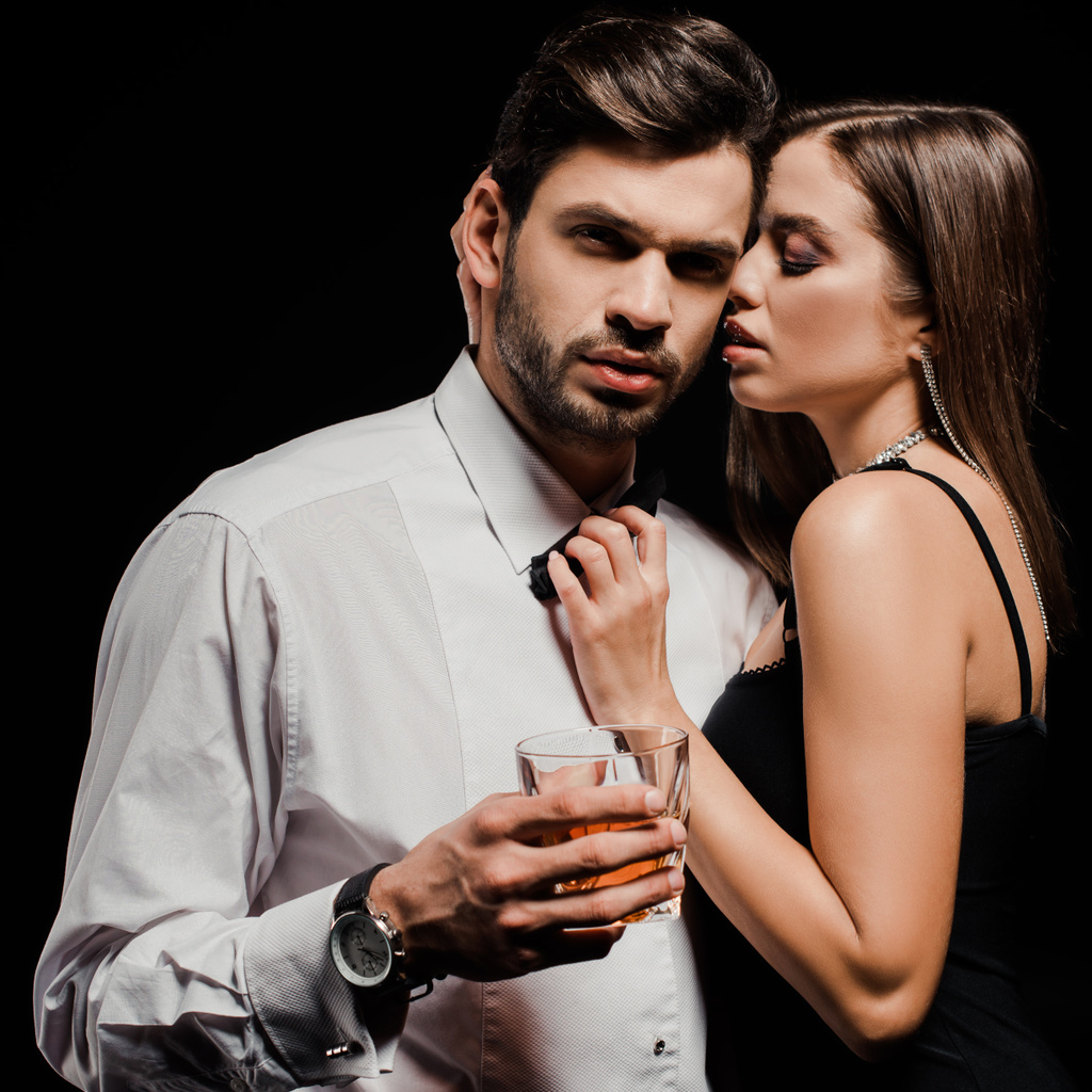 ελκυστική γυναίκα που αγγίζει παπιγιόν του όμορφου άνδρα με ένα ποτήρι ουίσκι απομονωμένο σε μαύρο  - Φωτογραφία, εικόνα