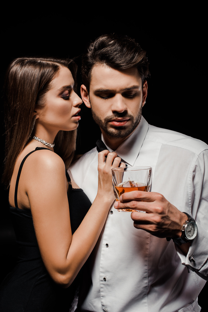соблазнительная женщина трогает галстук-бабочку красивого мужчины со стаканом виски, изолированного на черном
  - Фото, изображение