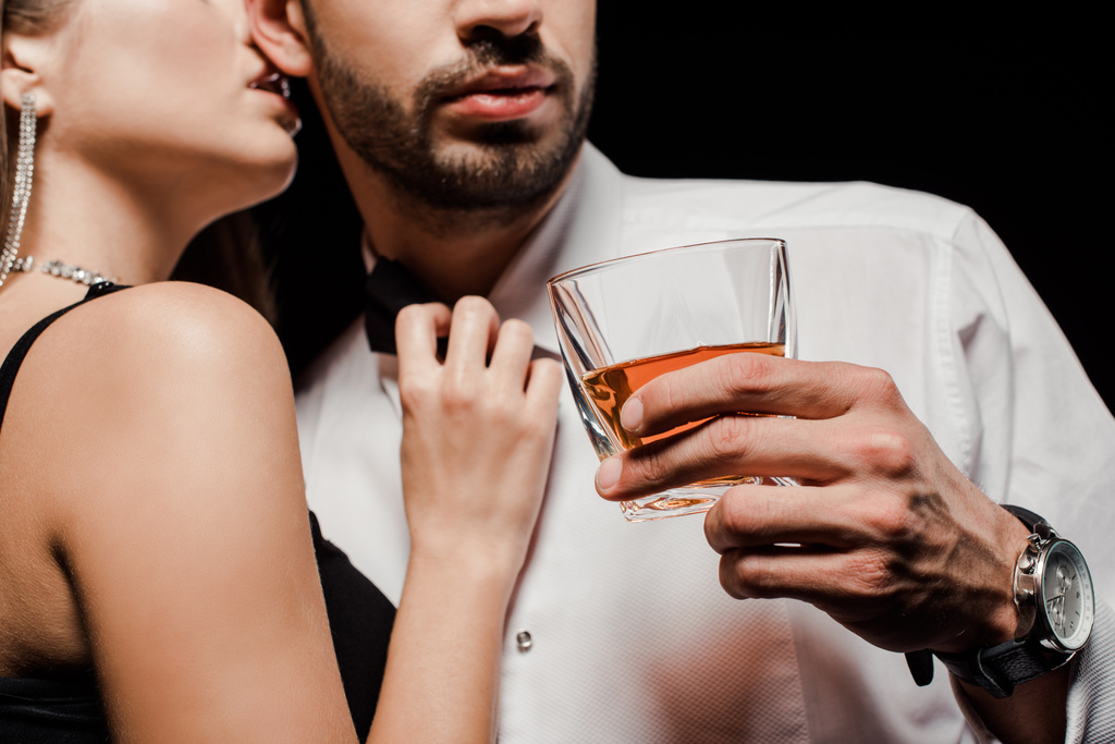 cropped view of woman touching bow tie and kissing man with glass of whiskey isolated on black  - Photo, Image