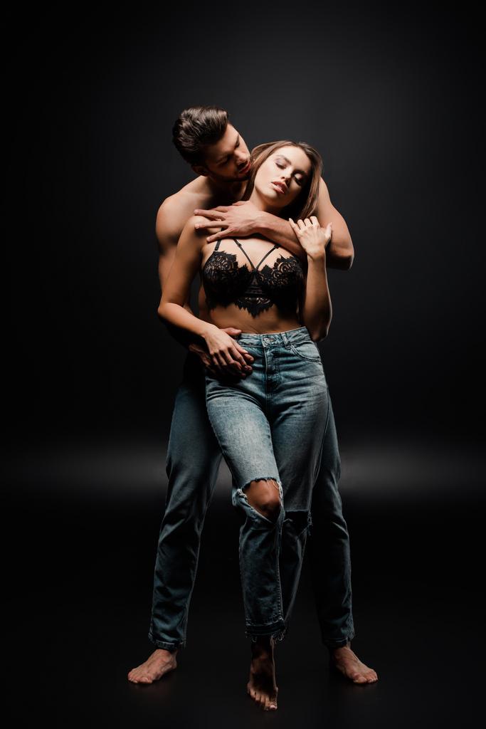 м'язистий чоловік обіймає привабливу дівчину в джинсах на чорному
  - Фото, зображення