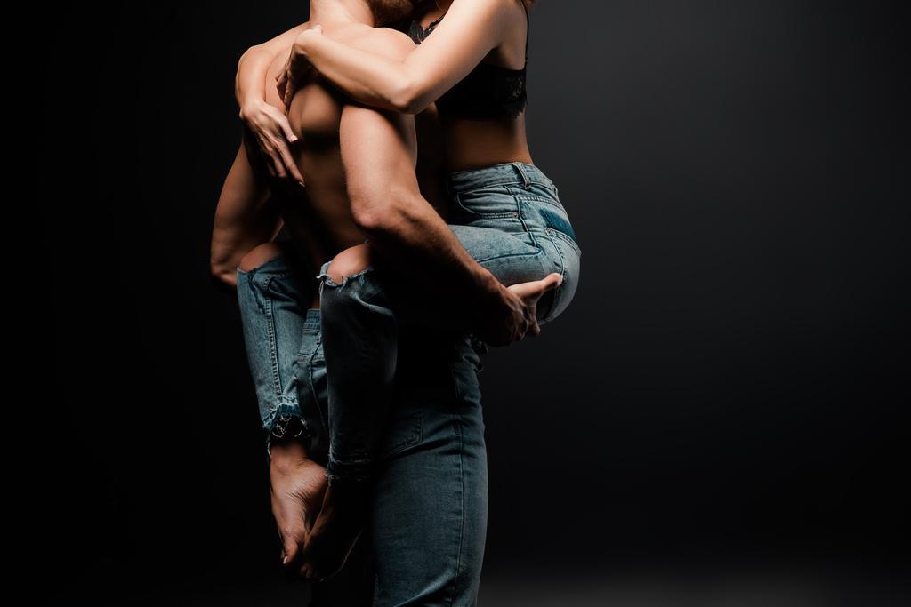 bijgesneden beeld van gespierde man houden in armen vrouw in spijkerbroek op zwart - Foto, afbeelding