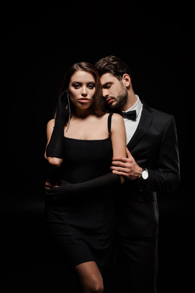 hombre guapo en traje abrazando hermosa mujer en guantes aislados en negro
  - Foto, imagen