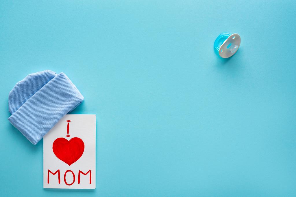 Top view of I love mom lettering on greeting card κοντά στο καπέλο και πιπίλα σε μπλε φόντο - Φωτογραφία, εικόνα