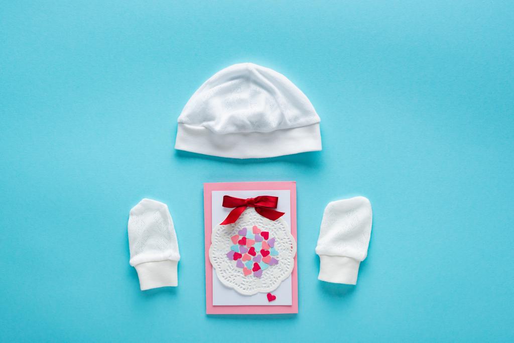 Top view of greeting card and baby hat and mittens on blue background, concept of mothers day  - Photo, Image