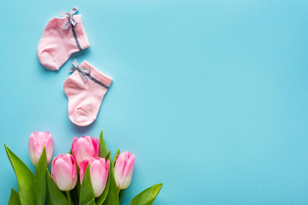 Bovenaanzicht van roze babysokken in de buurt van tulpen op blauw oppervlak, concept van moederdag  - Foto, afbeelding