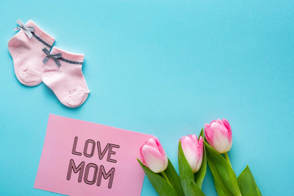Vista superior de meias de bebê rosa com tulipas e amor mãe lettering no cartão de saudação na superfície azul
 - Foto, Imagem