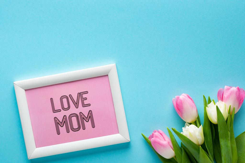 Top view of frame with i love mom lettering near bouquet of tulips on blue surface - Photo, Image