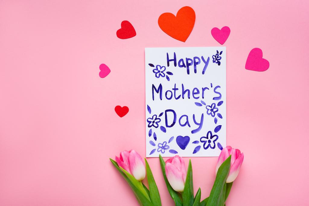 Top view of greeting card with happy mothers day lettering, tulips and paper hearts on pink background - Photo, Image