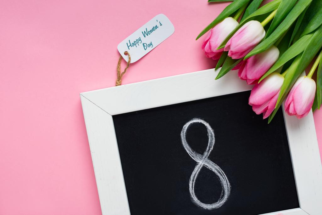 Top view of chalkboard with 8 number, paper label with happy womens day lettering and tulips on pink surface - Photo, Image
