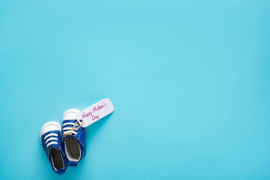 Vista dall'alto di stivaletti bambino con felice giorno delle madri lettering su etichetta di carta su sfondo blu
 - Foto, immagini