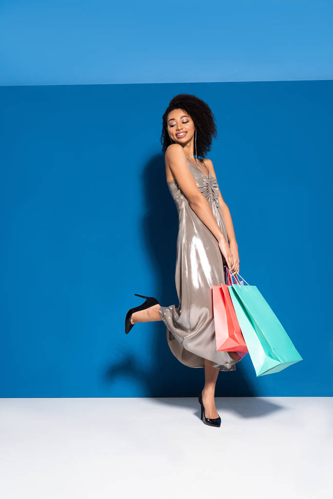 uśmiechnięta, piękna, afrykańska kobieta w srebrnej sukience, pozująca z torbami na zakupy na niebieskim tle - Zdjęcie, obraz