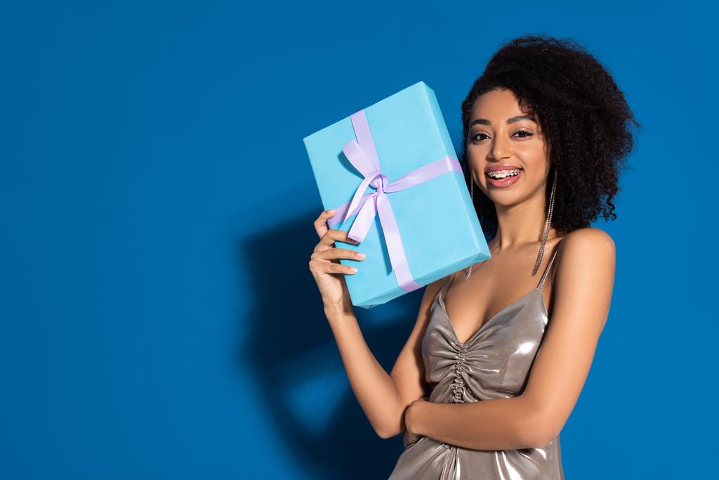 heureuse belle femme afro-américaine en robe d'argent tenant cadeau sur fond bleu
 - Photo, image
