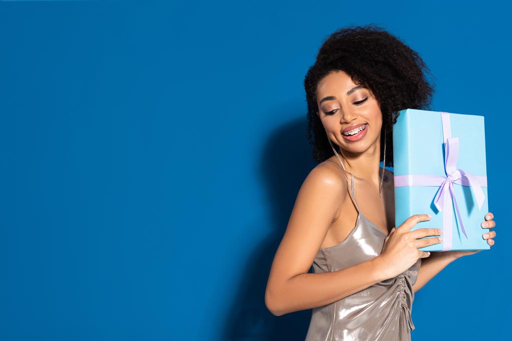 Gülümseyen, güzel, Afro-Amerikalı, gümüş elbiseli, elinde hediyesiyle mavi arka planda. - Fotoğraf, Görsel