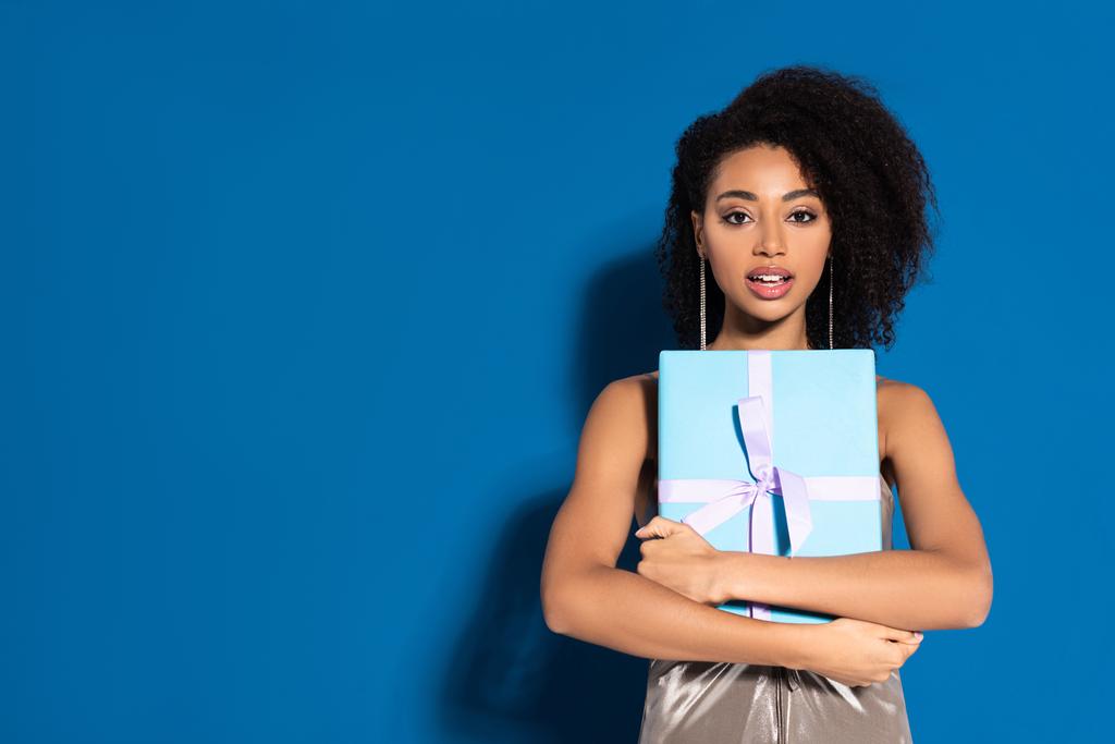 excited beautiful african american woman in silver dress with open mouth holding gift on blue background - Photo, Image