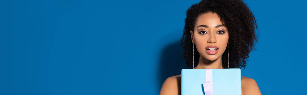 aufgeregt schöne afrikanisch-amerikanische Frau mit offenem Mund hält Geschenk auf blauem Hintergrund, Panoramaaufnahme - Foto, Bild