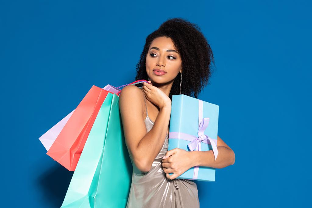 χαμογελαστή όμορφη Αφροαμερικανή γυναίκα σε ασημί φόρεμα κρατώντας κουτί δώρου και τσάντες ψώνια, ενώ κοιτάζοντας μακριά σε μπλε φόντο - Φωτογραφία, εικόνα