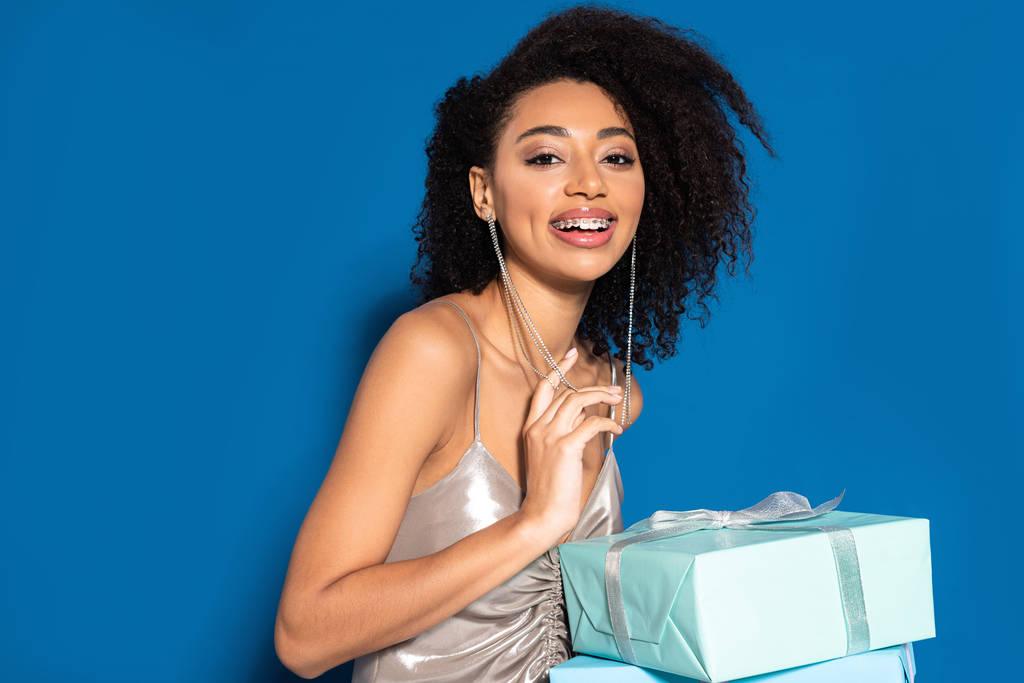 smiling beautiful african american woman in silver dress holding gifts on blue background - Fotoğraf, Görsel