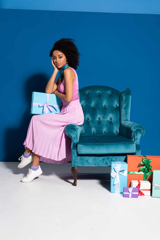 donna afroamericana seduta su una poltrona di velluto vicino a scatole regalo su sfondo blu
 - Foto, immagini