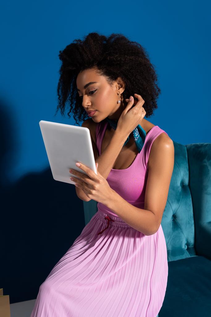Afroamerykanka siedząca w welurowym fotelu z cyfrową tabletką na niebieskim tle - Zdjęcie, obraz