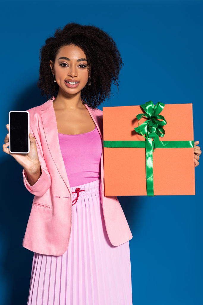élégante femme afro-américaine souriante avec cadeau montrant smartphone avec écran blanc sur fond bleu
 - Photo, image