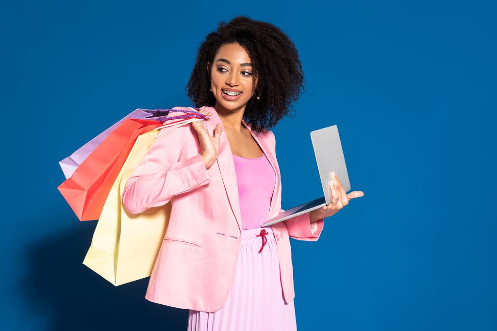 usmívající se elegantní africká americká obchodnice s nákupními taškami pomocí notebooku na modrém pozadí - Fotografie, Obrázek