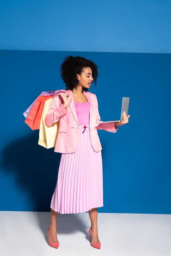 elegante afrikanisch-amerikanische Geschäftsfrau mit Einkaufstaschen mit Laptop auf blauem Hintergrund - Foto, Bild