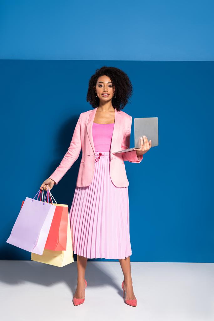 elegante mujer de negocios afroamericana con bolsas de compras utilizando el ordenador portátil sobre fondo azul
 - Foto, imagen