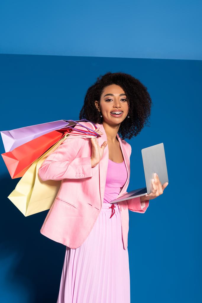souriant élégante femme d'affaires afro-américaine avec des sacs à provisions à l'aide d'un ordinateur portable sur fond bleu
 - Photo, image