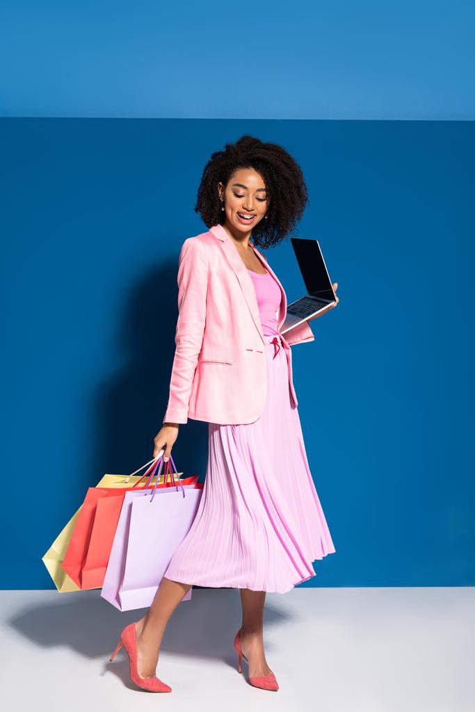 glimlachende elegante Afrikaanse Amerikaanse zakenvrouw met boodschappentassen met laptop op blauwe achtergrond - Foto, afbeelding