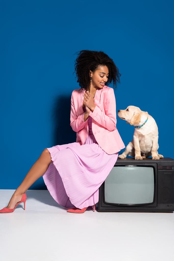 sonriente africana americana mujer sentado en vintage tv con golden retriever cachorro en azul fondo
 - Foto, imagen