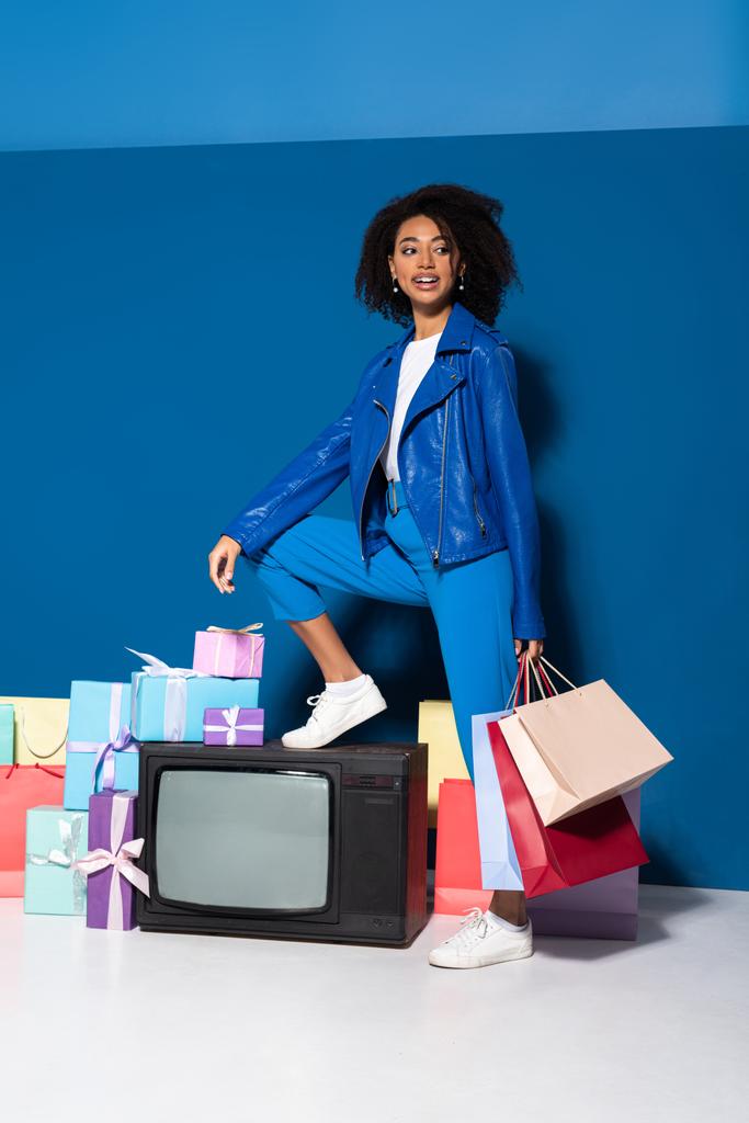 sonriente mujer afroamericana sentada en la televisión vintage con bolsas de compras sobre fondo azul
 - Foto, imagen
