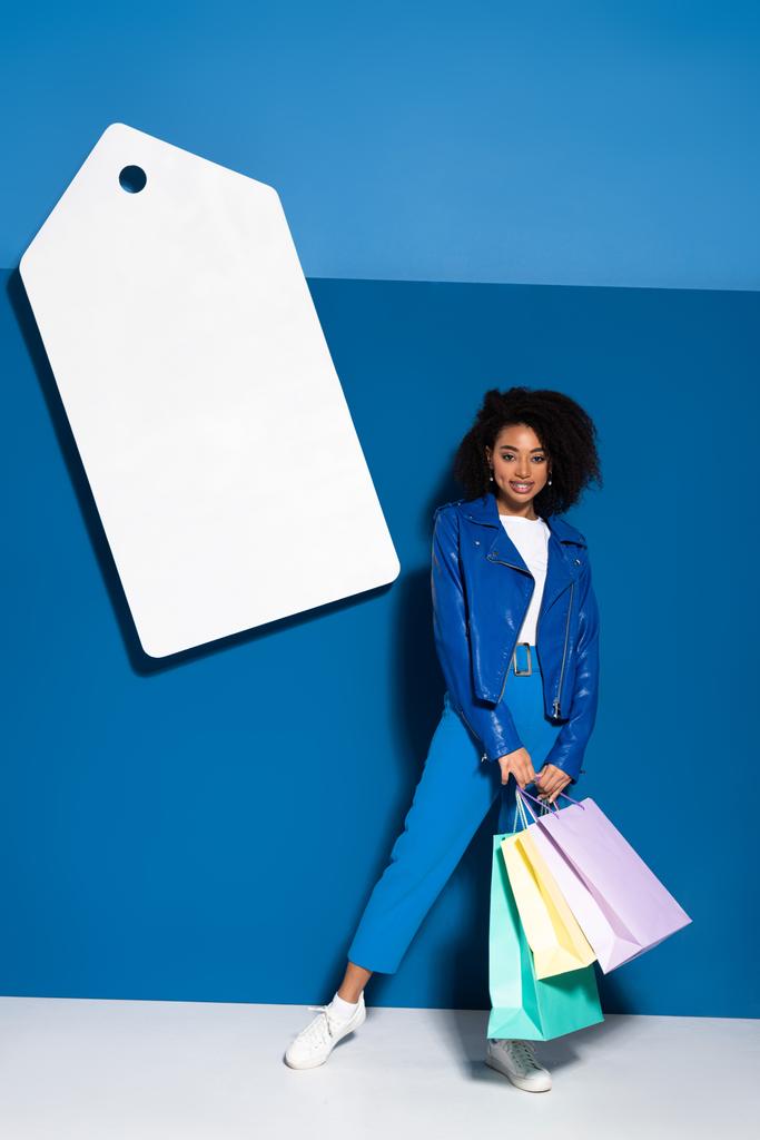 usmívající se africká americká žena s nákupními taškami v blízkosti velké prázdné cenové značky na modrém pozadí - Fotografie, Obrázek