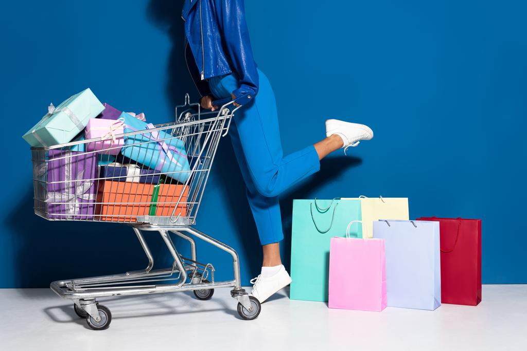 vista recortada de mujer afroamericana con carrito de compras lleno de regalos cerca de bolsas de compras sobre fondo azul
 - Foto, Imagen