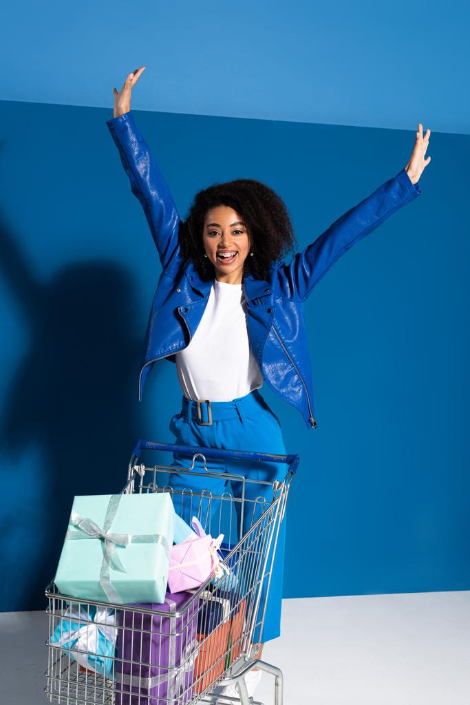 happy african american woman with hands in air and shopping cart full of gifts on blue background - Fotoğraf, Görsel