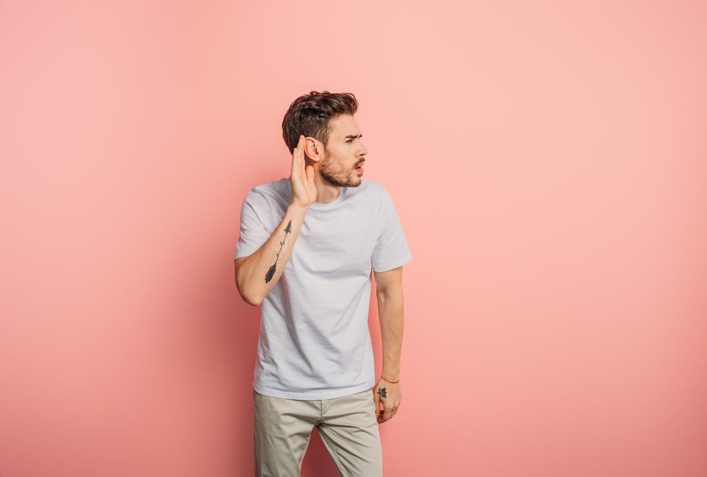 confused young man showing cant hear gesture by holding hand near ear on pink background - Photo, Image