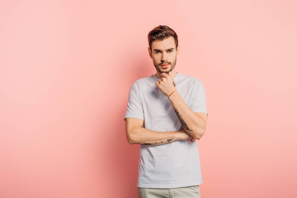 thoughtful young man touching chin while looking at camera on pink background - Photo, Image