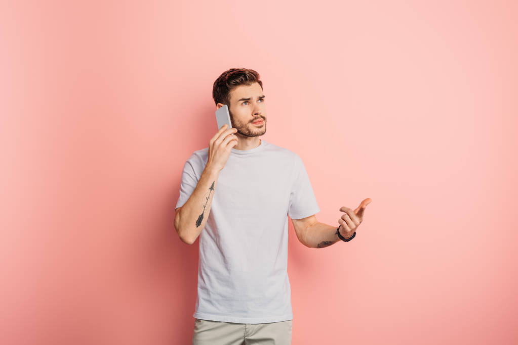 doordachte jonge man weg te kijken tijdens het praten op smartphone op roze achtergrond - Foto, afbeelding