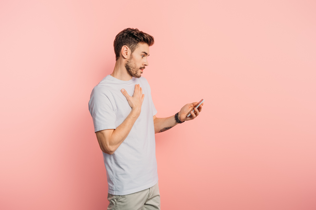 удивленный молодой человек трогает грудь, держа смартфон на розовом фоне
 - Фото, изображение