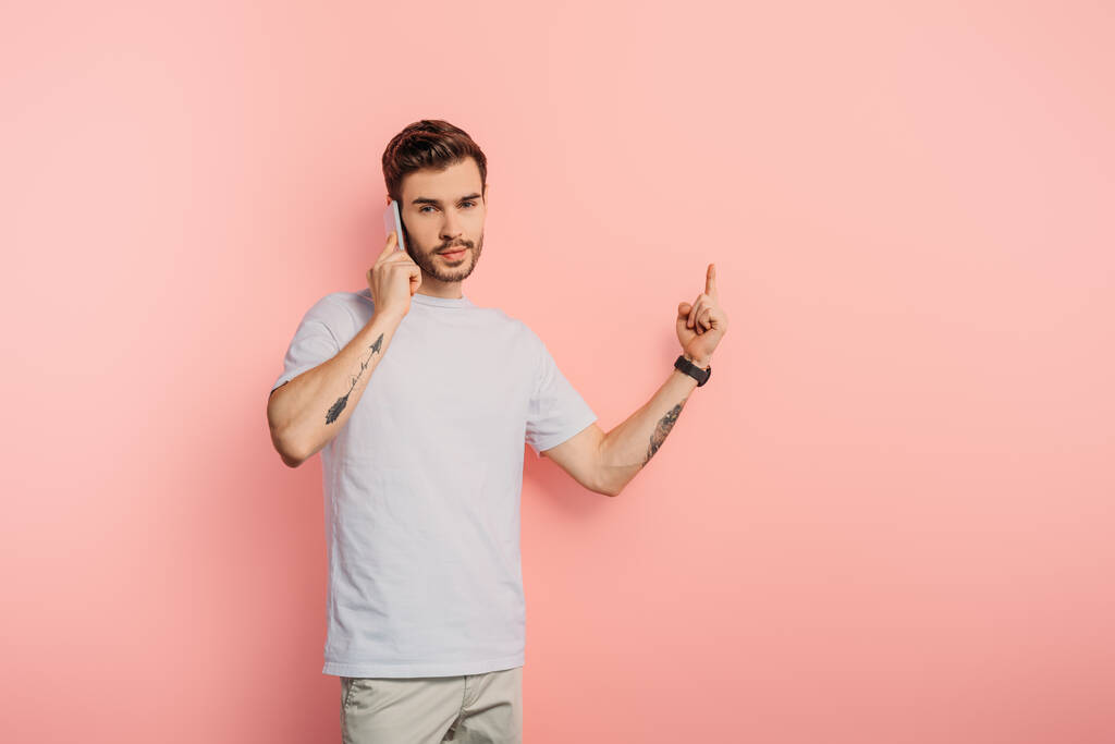 joven serio mostrando gesto de idea mientras habla en el teléfono inteligente sobre fondo rosa
 - Foto, Imagen