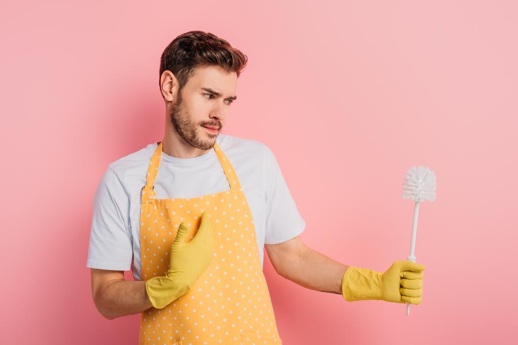 Verwirrter junger Mann in Schürze und Gummihandschuhen zeigt mit der Hand auf sich selbst, während er die Toilettenbürste auf rosa Hintergrund betrachtet - Foto, Bild