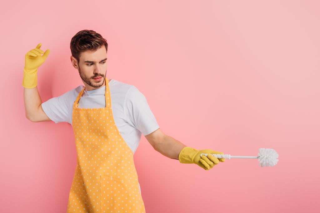 συμπυκνωμένος άνδρας με ποδιά και λαστιχένια γάντια που μιμείται περίφραξη με βούρτσα τουαλέτας σε ροζ φόντο - Φωτογραφία, εικόνα