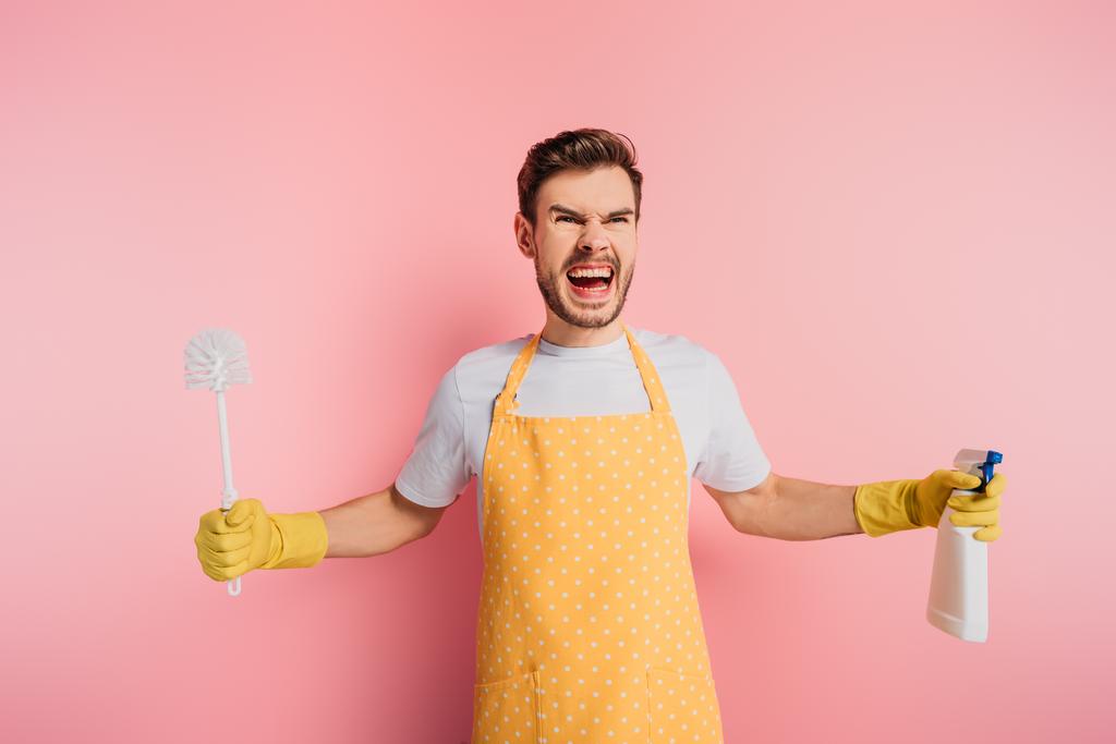 aggressive young man in apron and rubber gloves holding toilet brush and spray bottle on pink background - Photo, Image