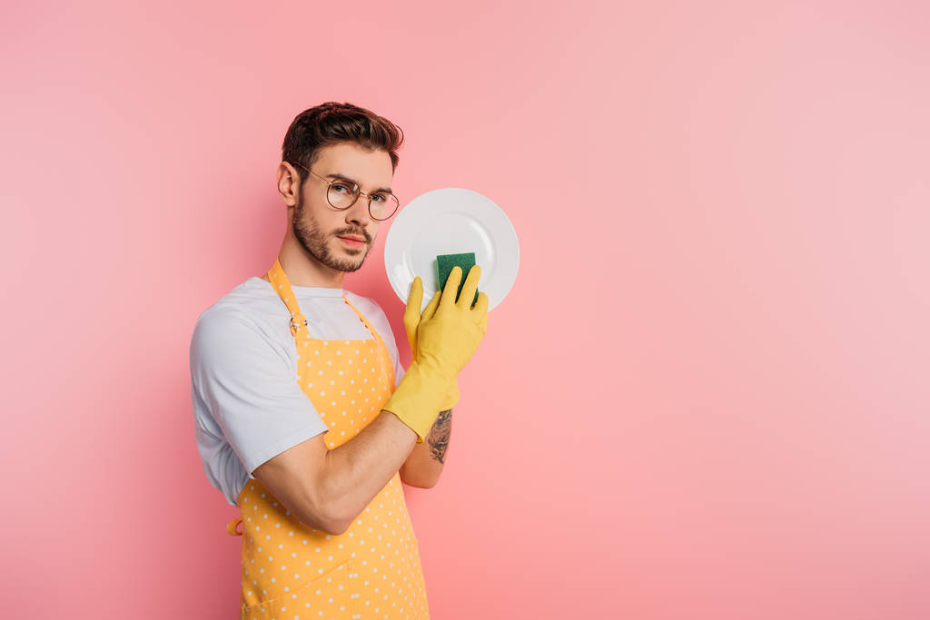 serious young man in apron and rubber gloves cleaning plate with sponge on pink background - Photo, Image