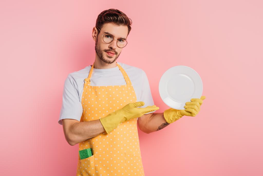 confident young man in apron and rubber gloves showing clean plate on pink background - Photo, Image