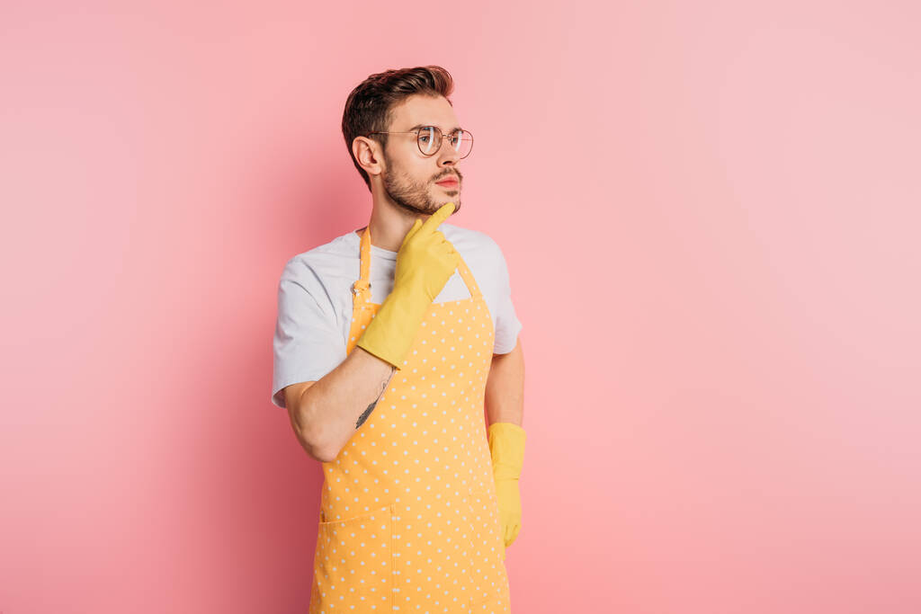 задумчивый молодой человек в фартуке и резиновых перчатках касаясь подбородка и глядя в сторону на розовый фон
 - Фото, изображение