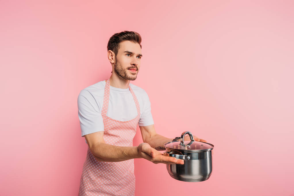 schöner junger Mann in Schürze imitiert Kellner, während er Kochtopf auf rosa Hintergrund hält  - Foto, Bild