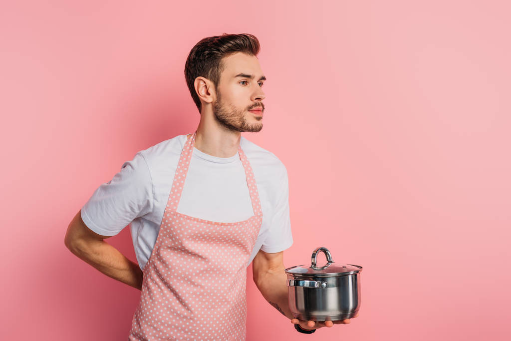 knappe jongeman in schort imiteren ober terwijl het houden van steelpan op roze achtergrond  - Foto, afbeelding