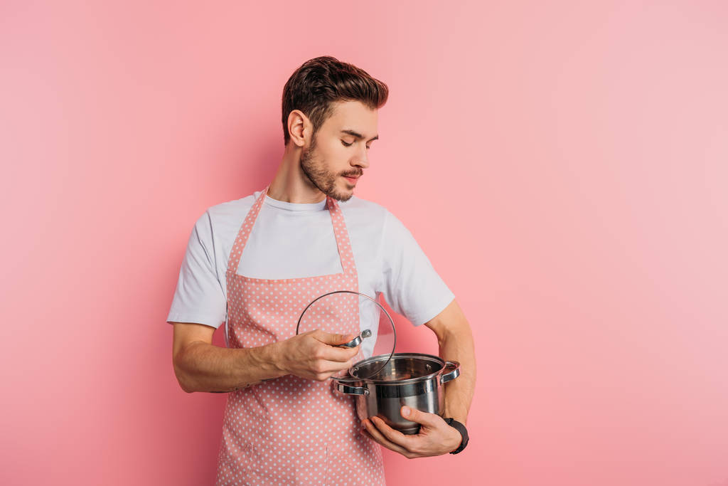 sérieux jeune homme dans tablier ouverture et regarder dans la casserole sur fond rose
  - Photo, image